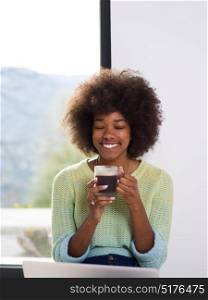 Young african american woman smiling sitting on the floor near bright window while looking at open laptop computer and holding mug at home