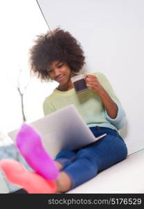 Young african american woman smiling sitting on the floor near bright window while looking at open laptop computer and holding mug at home