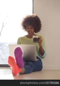 Young african american woman smiling sitting on the floor near bright window while looking at open laptop computer and holding mug at home