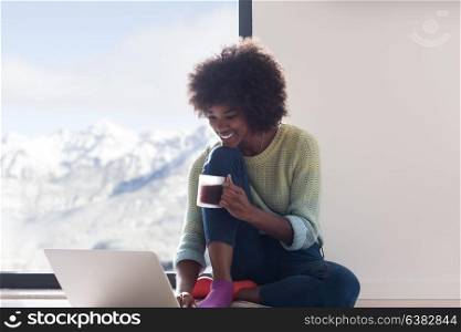 Young african american woman smiling sitting on the floor near bright window while looking at open laptop computer and holding mug at home