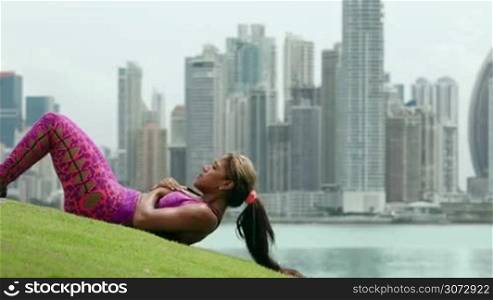 Young african american woman training abdominals and exercising early morning in the city. She trains on grass along the Cinta Costera in Panama City. Long shot