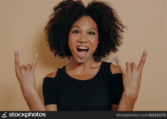 Young african american woman wearing black tshirt shouting with crazy facial expression doing rock-n-roll symbol with hands up likeμsic star, isolated over bei≥studio background with©space. Young woman wearing black tshirt shouting with crazy facial expression doing rock-n-roll symbol