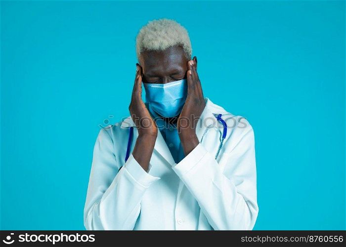 Young african doctor man in professional medical white coat having headache, studio portrait. Guy putting hands on head, isolated on blue background. Concept of problems, medicine, illness.. Young african doctor man in professional medical white coat having headache, studio portrait. Guy putting hands on head, isolated on blue background. Concept of problems, medicine, illness