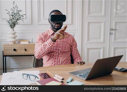 Young African ethnicity man entrepreneur in VR headset glasses immersed in 3D gaming in virtual reality relaxing during remote working online sitting at desk at modern home office in front of laptop. Young African ethnicity man entrepreneur in VR headset glasses immersed in 3D gaming