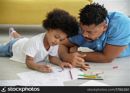 Young African father and little boy lying on floor and drawing coloring on book together with color pencil crayon at home