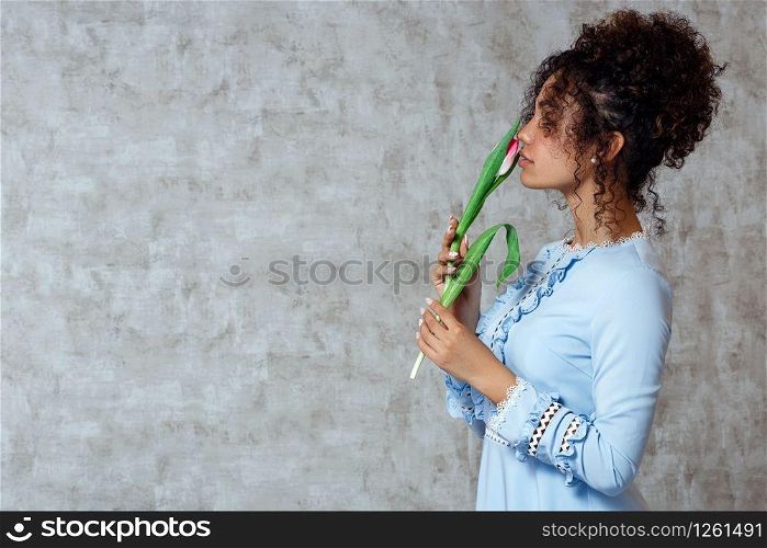 Young African girl in a blue dress with a Tulip on a gray background. The concept of women&rsquo;s day and spring