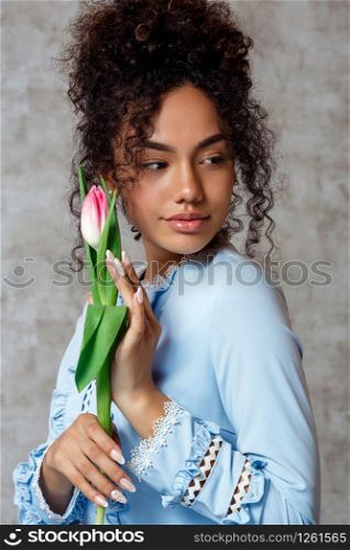 Young African girl in a blue dress with a Tulip on a gray background. The concept of women&rsquo;s day and spring