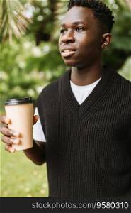 Young African man holding disposable brown paper coffee cup on street outdoors, smiling. Coffee or tea to go mockup. Young African man holding disposable paper coffee cup on street