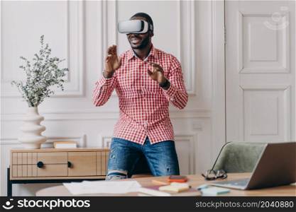 Young african man in VR headset interacting with digital interface while testing and using innovative device for business, standing in modern office in virtual reality helmet. Cyberspace experience. African man in VR headset interacting with digital interface, using innovative device for business