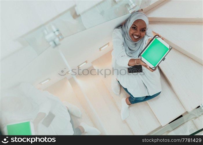 Young African modern Muslim woman using tablet computer while sitting on the stairs at home wearing hijab clothes top view. High-quality photo. Young african modern muslim woman using tablet computer while sitting on the stairs at home wearing hijab clothes top view