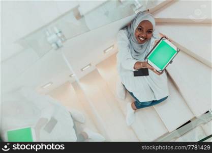 Young African modern Muslim woman using tablet computer while sitting on the stairs at home wearing hijab clothes top view. High-quality photo. Young african modern muslim woman using tablet computer while sitting on the stairs at home wearing hijab clothes top view
