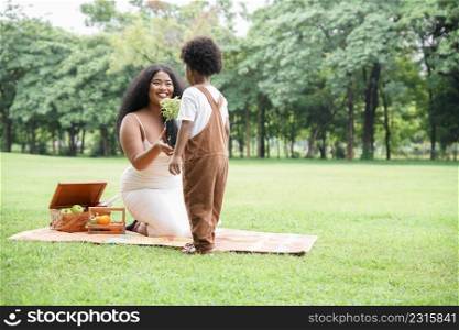 Young African mother smiling and giving little flowers tree to her son to plant at garden. African family picnic together sitting on mat at park with basket of fruits and drinks in glass bottle