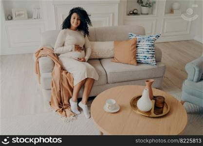 Young african pregnant woman in casual dress caressing her belly while sitting on cozy couch in stylish living room ambiance. Happy future mom expecting baby and maternity leisure time at home concept. Young african pregnant woman in casual dress caressing her belly while sitting on couch at home