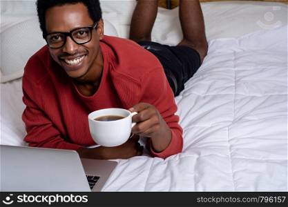 Young afro american man looking at camera and using his laptop with cup of coffee in bed.