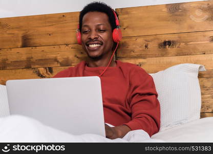 Young afro american man using his laptop with headphones in bed.