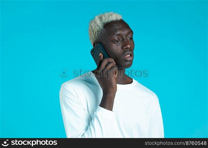 Young afro man speaks serious on phone. Guy holding and using smart phone. Blue studio background. Young afro man speaks serious on phone. Guy holding and using smart phone. Blue studio background.