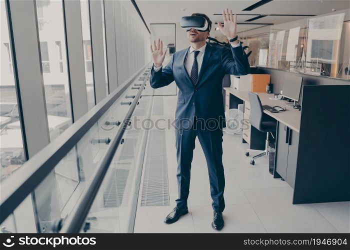 Young amazed businessman in VR headset trying to touch something with hands while standing in modern office, being excited while exploring virtual reality at work, dressed in formal clothes. Young amazed businessman in VR headset trying to touch something with hands while standing in modern office