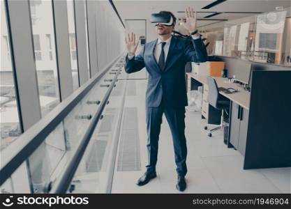 Young amazed businessman in VR headset trying to touch something with hands while standing in modern office, being excited while exploring virtual reality at work, dressed in formal clothes. Young amazed businessman in VR headset trying to touch something with hands while standing in modern office