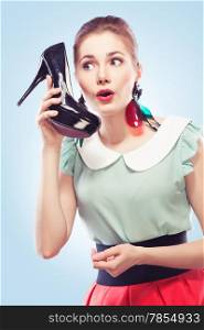 Young amazed woman using a shoe like a telephone holding it near her face and talking, blue background. Pin-up style.