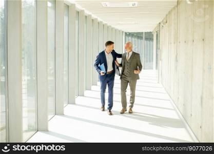 Young and a senior businessman walk down an office hallway, deep in conversation. They are both dressed professionally, reflecting their business acumen and status. They are navigating the complexities of the corporate world, finding solutions and making decisions together