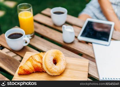 Young And Attractive Woman Having Morning Breakfast In Green Garden With French Croissant, Donuts, Coffee Cup, Orange Juice, Tablet and Notes Book On Wooden Table