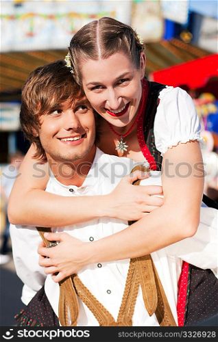 Young and beautiful couple in traditional Bavarian Tracht - Dirndl and Lederhosen - embracing each other on a fair like a Dult or the Oktoberfest; both are standing in front of a typical booth