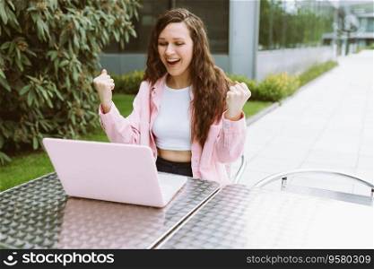 Young and confident woman in casual clothes smile, raise her hands and celebrating work success in office while sitting and working on her laptop in street cafe restaurant on summer day. Freelance, online business, online work, win, luck, self-employment. Copy space. Young woman raise her hands and celebrating win, sitting and working on her laptop in street cafe