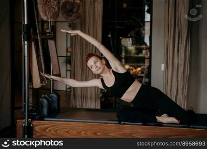 Young and fit ginger woman exercising and stretching on pilates cadillac machine with arms outstretched to side, showing flexibility and relaxing pose. Concept of fitness and wellbeing concept. Young female pilates instructor exercising and stretching on cadillac reformer