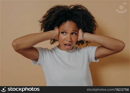 Young annoyed african woman covering her ears and keeping mouth opened, feeling stressed, does not want to hear you while standing isolated on beige background, unhappy female suffering from headache. Young annoyed african woman covering her ears and keeping mouth opened isolated on beige background