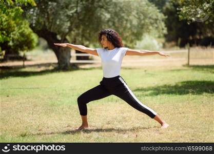 Young Arab woman doing yoga in nature. North African female wearing sport clothes doing Warrior II figure in urban park.