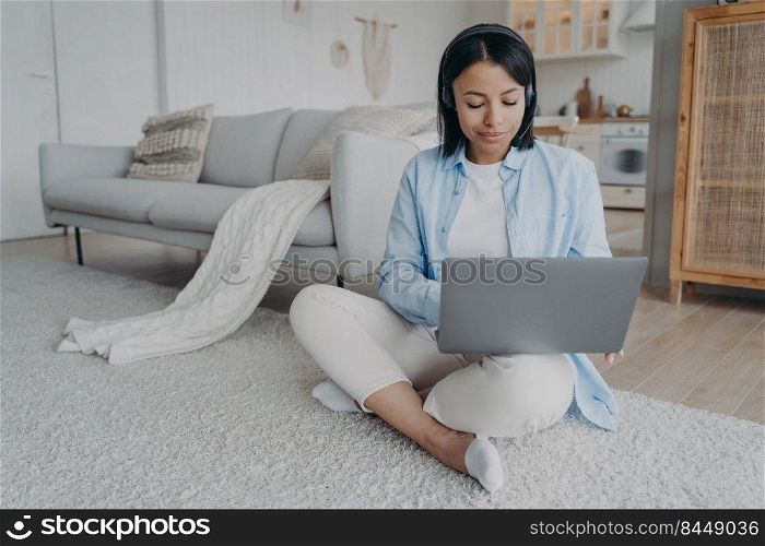 Young arab woman in headset and mic is sitting on floor having video chat on laptop. European girl is smiling and tyπng on pc. Freelancer has∫er≠t session. Student has virtual≤sson.. Young woman is sitting on floor having video chat on laptop. Girl is smiling and tyπng on pc.