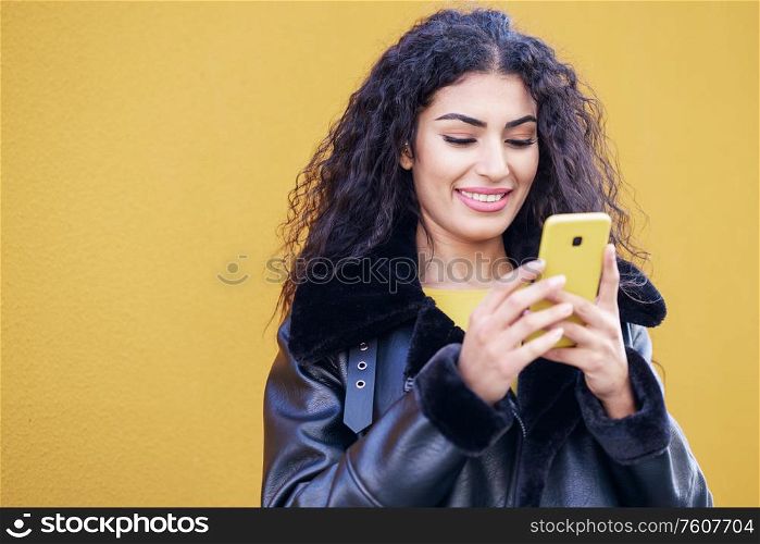 Young Arab woman texting a text message with her smartphone against yellow urban wall.. Young Arab woman walking in the street using her smartphone