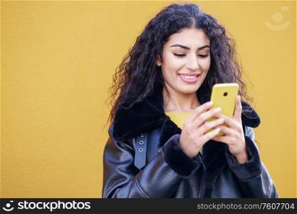 Young Arab woman texting a text message with her smartphone against yellow urban wall.. Young Arab woman walking in the street using her smartphone