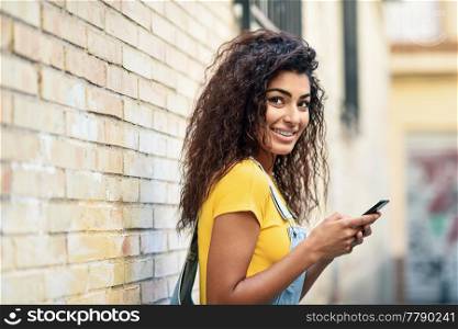 Young Arab woman texting with her smart phone on brick urban wall. Smiling North Afrincan girl in casual clothes with black curly hairstyle.