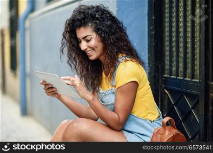 Young Arab woman using her digital tablet outdoors. Smiling North African female in casual clothes with black curly hairstyle.