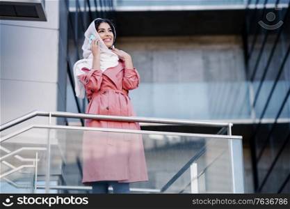 Young Arab woman walking on the street wearing hijab headscarf using smart phone in a office building. Business background.. Young Muslim woman wearing hijab using her smartphone.