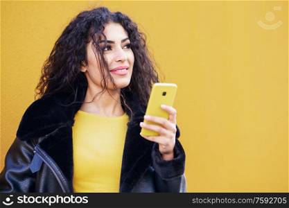 Young Arab woman wearing casual clothes,recording voice note with her smartphone against yellow urban wall.. Arab woman walking in the street using her smartphone