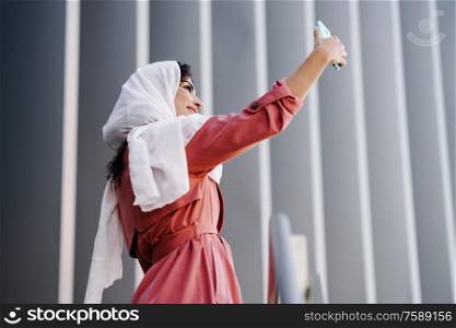 Young Arab Woman wearing hijab headscarf photograph with a smartphone outdoors.. Arab Woman with hijab taking selfie with smartphone