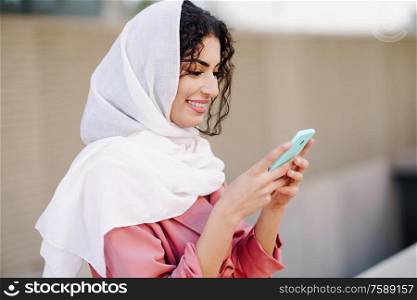 Young Arab woman wearing hijab headscarf texting message with her smartphone in urban background.. Young Muslim woman wearing hijab texting message with her smartphone.