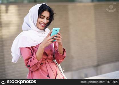 Young Arab woman wearing hijab headscarf texting message with her smartphone in urban background.. Young Arab woman wearing hijab texting message with her smartphone.