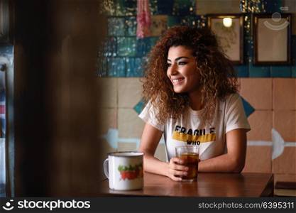 Young arabic smiling woman with black curly hairstyle sitting in a beautiful bar with vintage decoration. Arab girl in casual clothes drinking a soda.