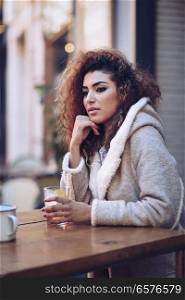 Young arabic woman sitting in an urban bar in the street. Arab girl in winter casual clothes drinking a soda outdoors.