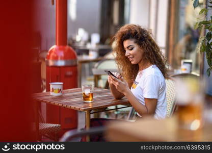 Young arabic woman smiling and sitting in an urban bar in the street looking at her smartphone. Arab girl in casual clothes drinking a soda outdoors.