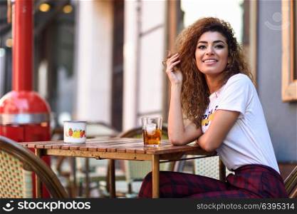 Young arabic woman smiling and sitting in an urban bar in the street. Arab girl in casual clothes drinking a soda outdoors.
