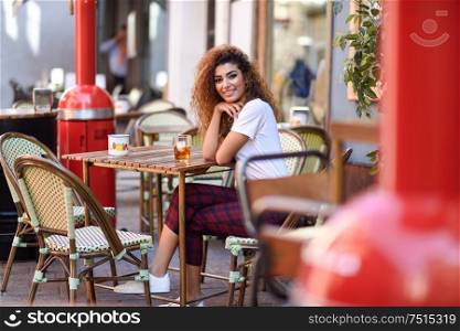Young arabic woman smiling and sitting in an urban bar in the street. Arab girl in casual clothes drinking a soda outdoors.. Arab girl in casual clothes drinking a soda in an outdoors bar