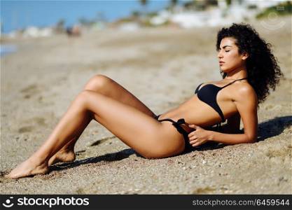 Young arabic woman with beautiful body in swimwear lying on the beach sand. Brunette female with curly long hairstyle wearing black bikini with eyes closed.