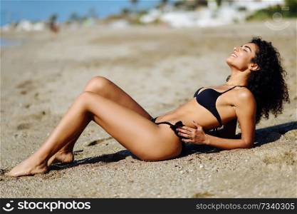 Young arabic woman with beautiful body in swimwear lying on the beach sand. Smiling female with curly long hairstyle wearing black bikini.. Arabic woman with beautiful body in bikini lying on the beach sand