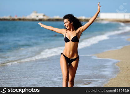Young arabic woman with beautiful body in swimwear on a tropical beach with open arms. Brunette female smiling with curly long hairstyle wearing black bikini.