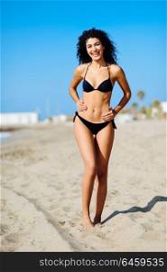 Young arabic woman with beautiful body in swimwear smiling in a tropical beach. Brunette female with curly long hairstyle wearing black bikini.