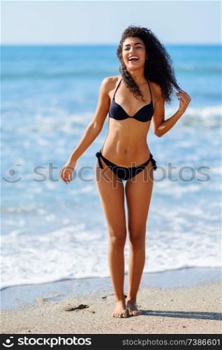 Young arabic woman with beautiful body in swimwear smiling in a tropical beach. Brunette female with curly long hairstyle wearing black bikini.. Young arabic woman with beautiful body in swimwear smiling on a tropical beach.